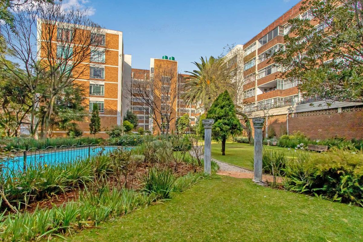 A view of the apartment complex grounds featuring a swimming pool, landscaped garden areas, and mature trees between the brick residential buildings.