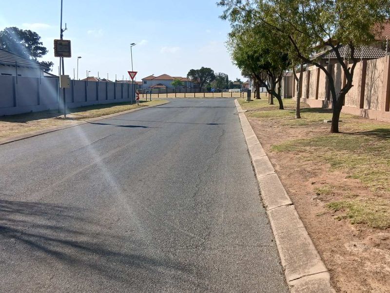 A view of the residential street approach featuring asphalt paving, concrete curbing, and perimeter boundary walls with electric fencing.