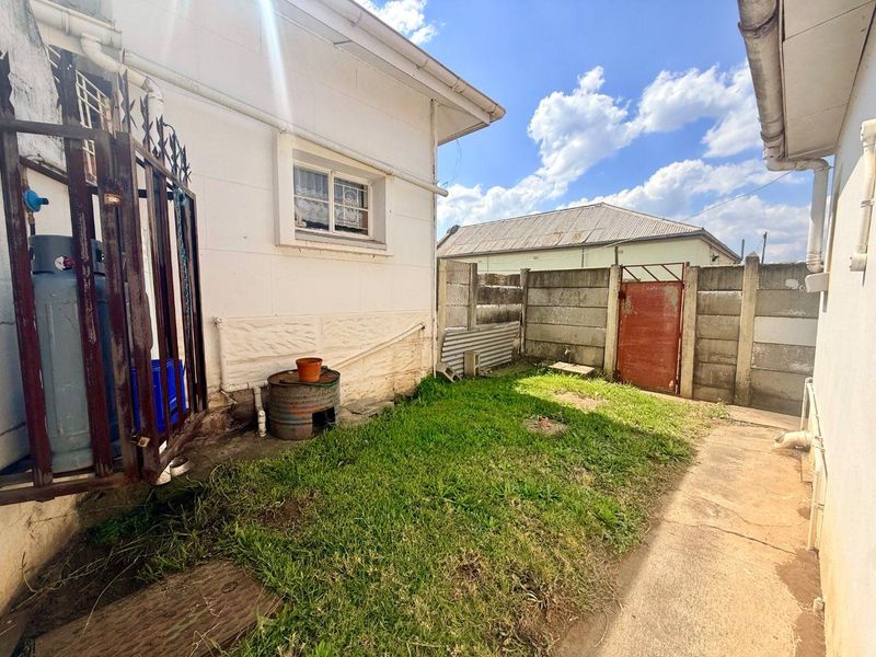 A rear exterior view of the property featuring a small lawn area, concrete paving, and a precast concrete boundary wall with a metal gate.