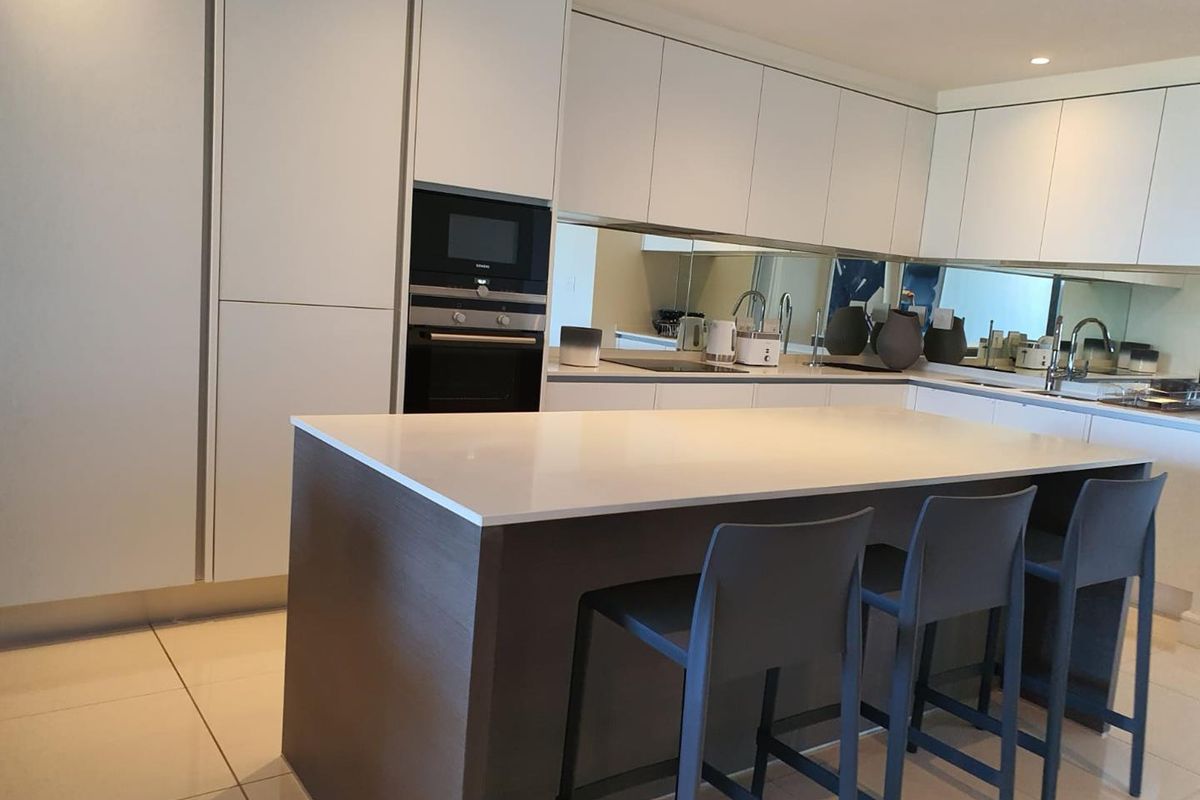 Modern kitchen featuring sleek white cabinetry, a central island with stone countertop, integrated oven, microwave, and a mirrored backsplash.