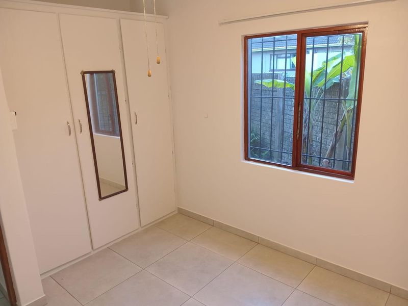 A clean, empty bedroom featuring neutral tiled flooring, built-in cupboards with a mirror, and a window fitted with security bars.