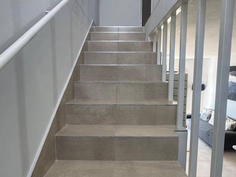 A bright staircase featuring modern neutral floor tiles, a white metal handrail, and a matching vertical balustrade leading to the upper level.
