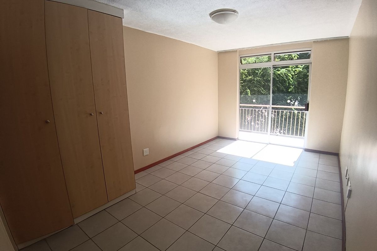 A bright, empty bedroom featuring neutral tiled flooring, built-in wooden cupboards, and a sliding glass door providing access to a balcony.
