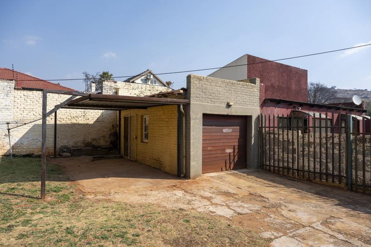 Rear exterior view featuring a single garage, attached metal carport, and concrete driveway area with surrounding brick boundary walls.
