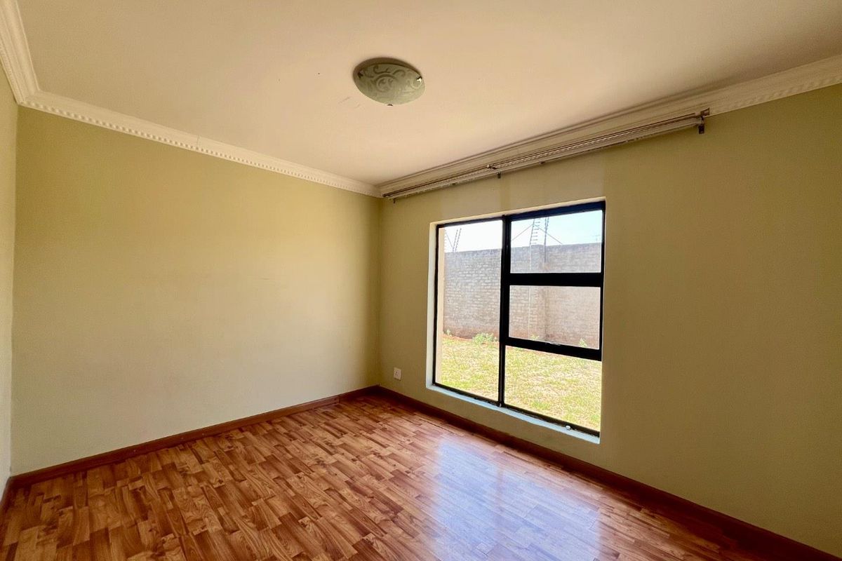 A bright, empty bedroom featuring wood-look laminate flooring, crown molding, and a large window overlooking the rear garden area.
