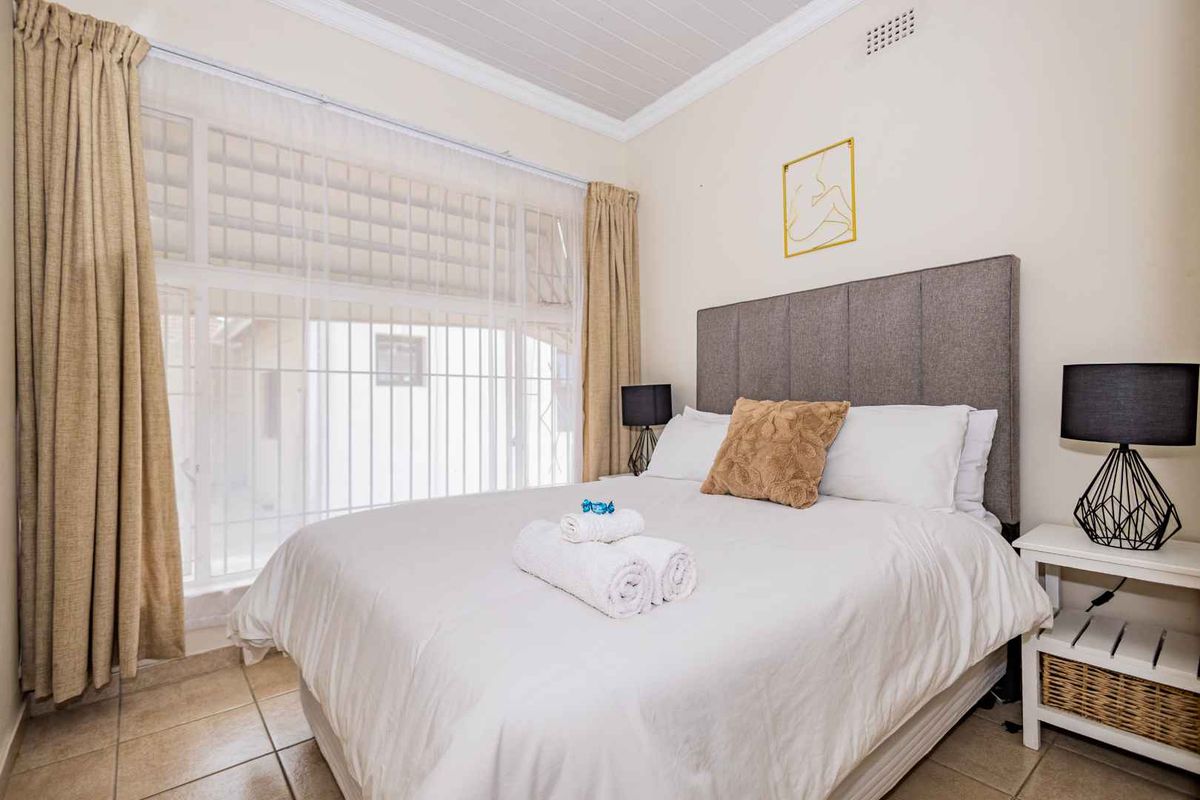 A well-lit bedroom featuring tiled flooring, a large window with security bars, and a panelled ceiling.