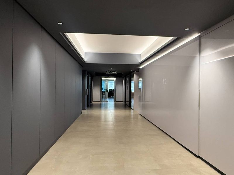 A modern, well-lit office hallway featuring sleek wall paneling, recessed ceiling lighting, and neutral tiled flooring.