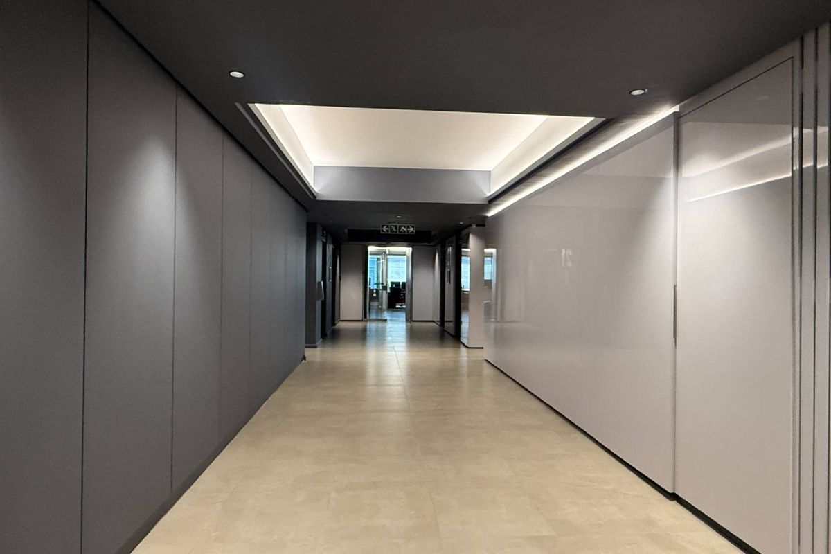 A modern, well-lit office hallway featuring sleek wall paneling, recessed ceiling lighting, and neutral tiled flooring.