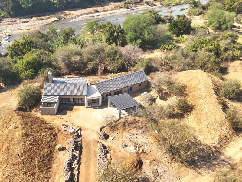 Aerial view of a modern house with a metal roof, situated in a natural landscape near a river, featuring a driveway and carport.