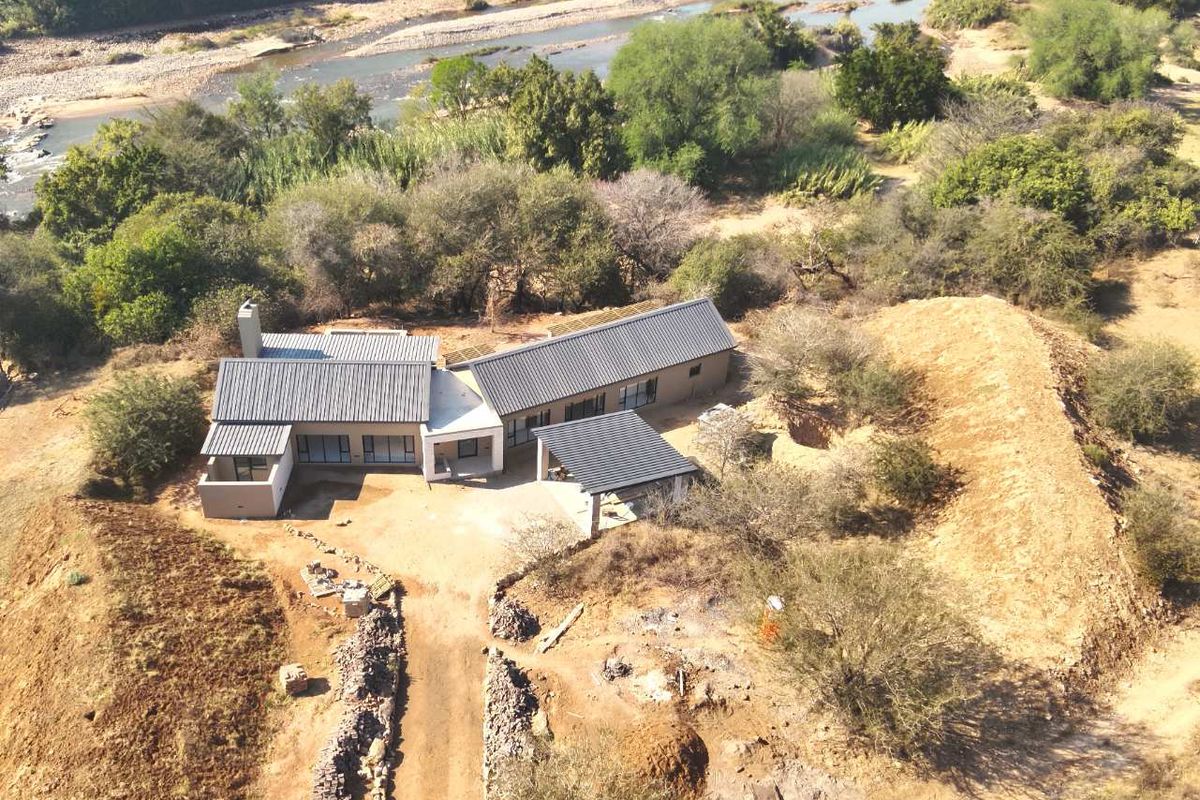 Aerial view of a modern house with a metal roof, situated in a natural landscape near a river, featuring a driveway and carport.