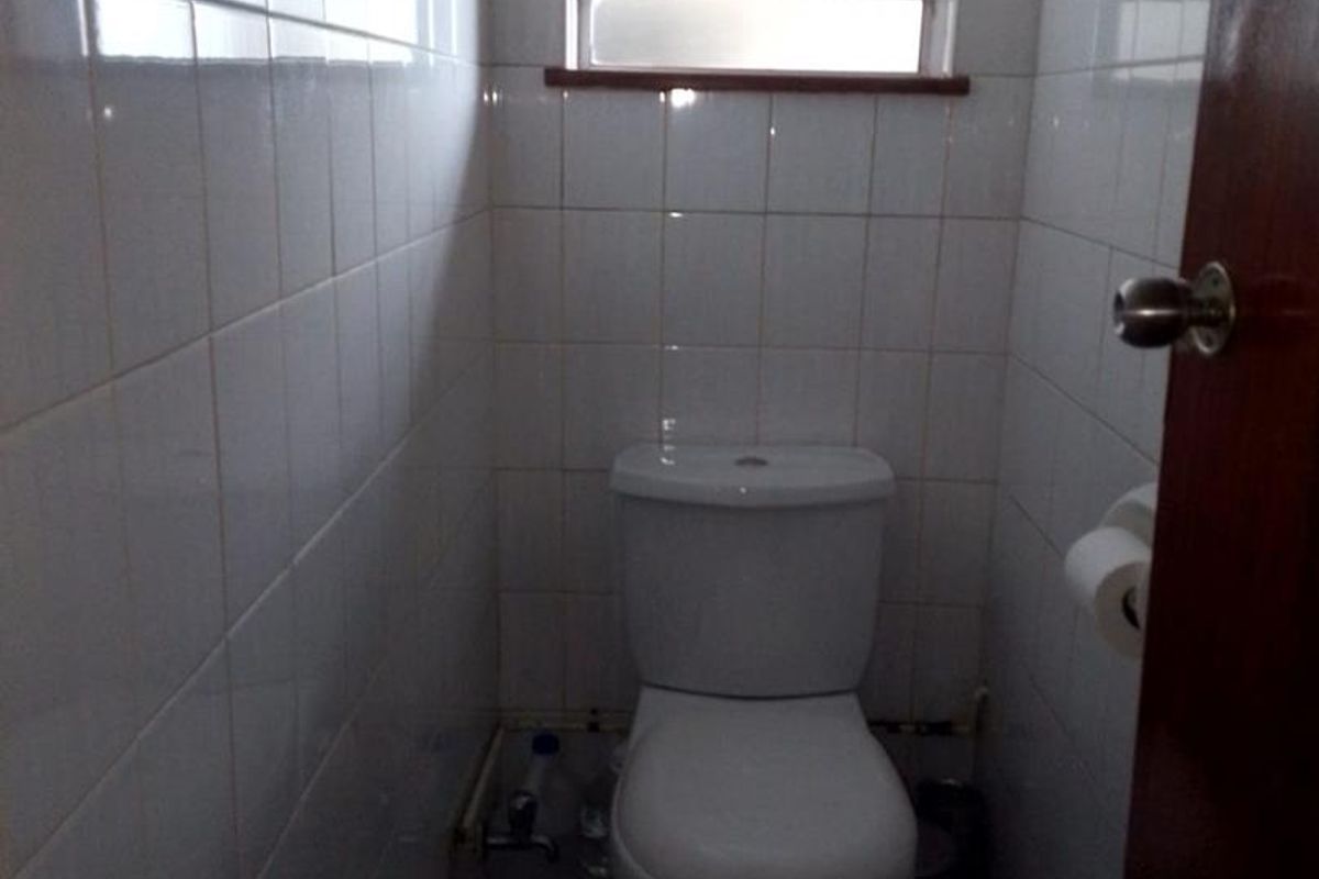 A compact, functional bathroom featuring white wall tiles, a standard toilet, and a window with security bars.