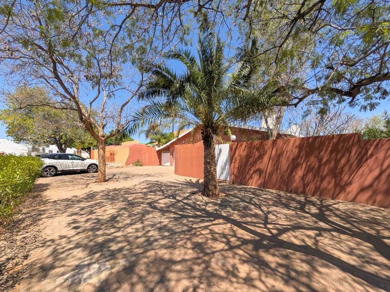 A view of the property's front exterior, featuring a paved driveway, mature landscaping, and a prominent boundary wall.