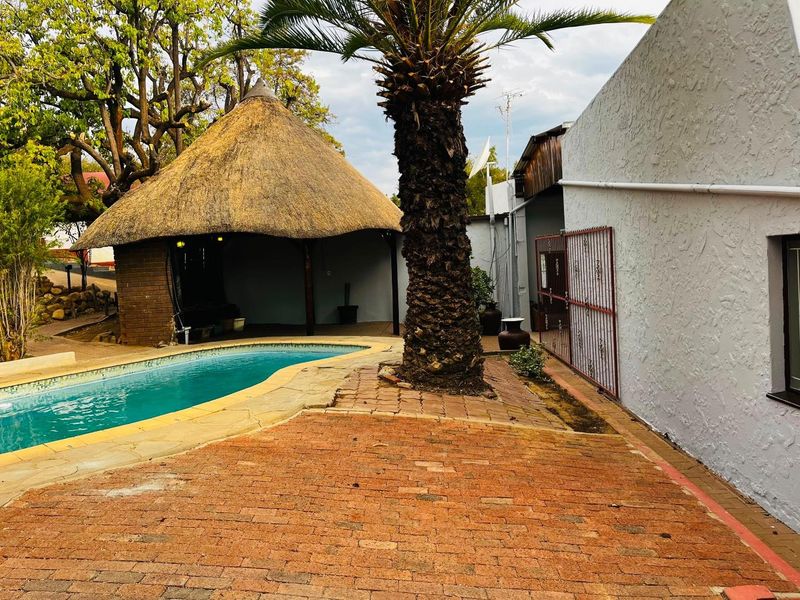 Outdoor pool area featuring a traditional thatch lapa, brick paved patio, and established palm tree, providing a relaxed space for outdoor entertainment.
