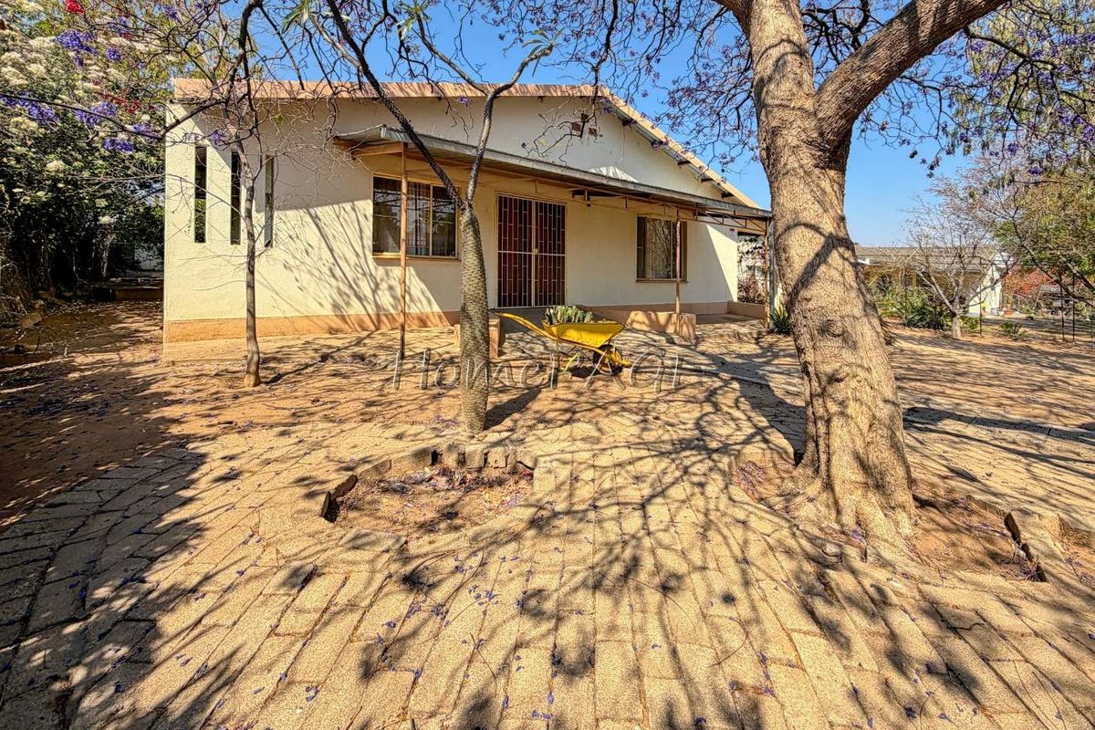 Rear view of the house featuring a paved patio area, mature trees, and security gates on the windows and entrance.