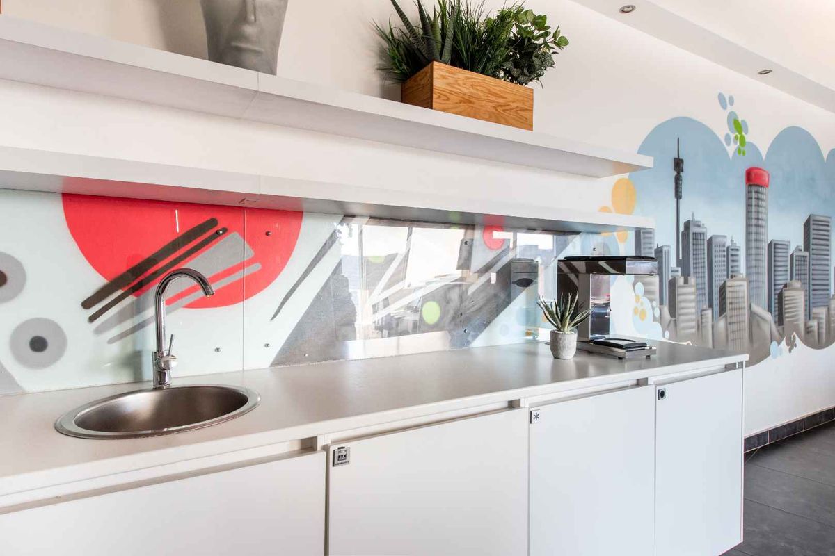 A modern office kitchenette featuring white cabinetry, a stainless steel sink, and a decorative glass splashback with a city-themed mural.