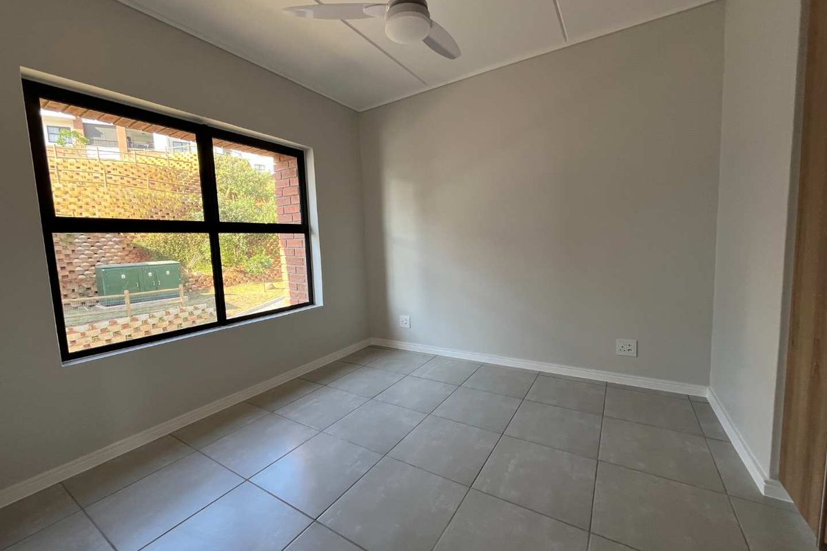 A bright, empty bedroom featuring neutral tiled flooring, a modern ceiling fan, and a large window with a view of the exterior landscape.
