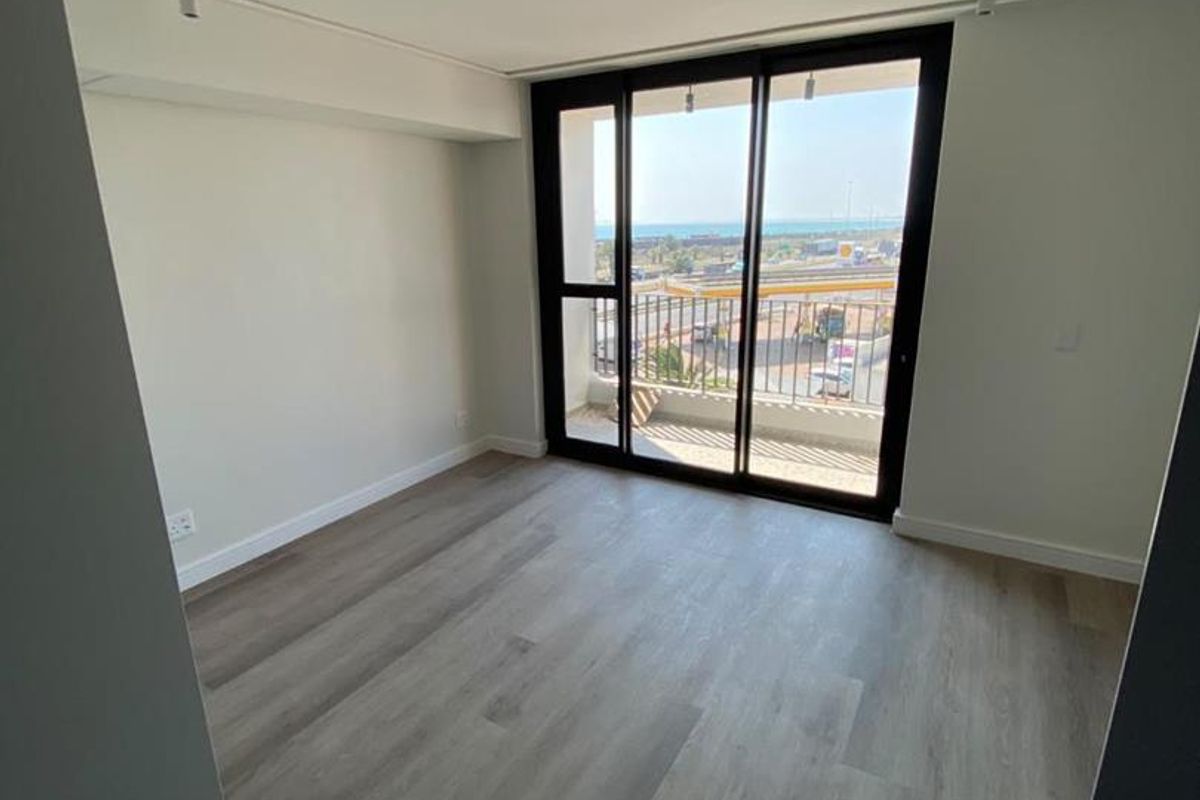 A bright, empty bedroom featuring modern laminate flooring, track lighting, and a large sliding glass door providing access to a balcony with coastal views.