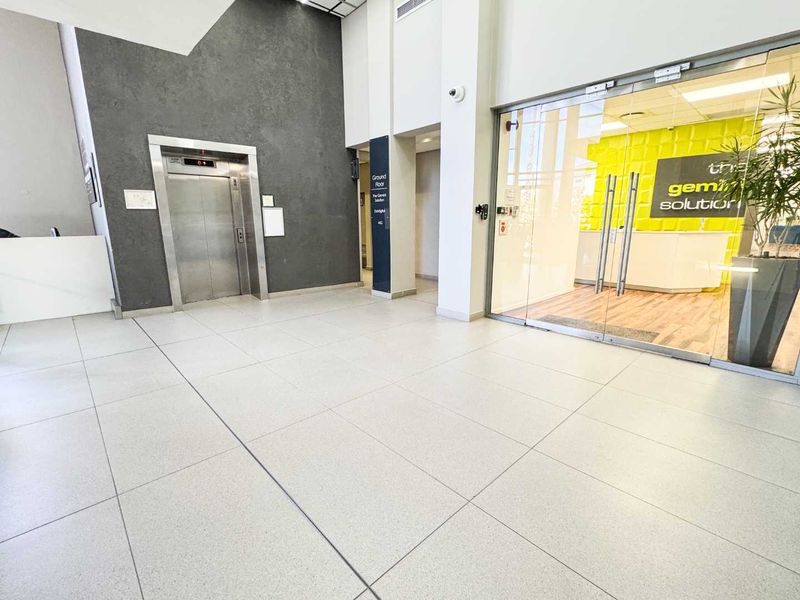 Modern commercial building lobby featuring tiled flooring, a stainless steel elevator, and a glass-enclosed office entrance.