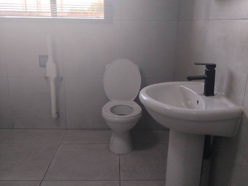 A clean, modern bathroom featuring a pedestal basin, toilet, and neutral wall and floor tiling, complemented by a wall-mounted storage cabinet.