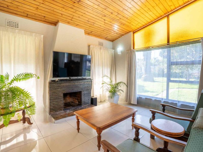 A bright living room featuring a brick fireplace, timber-clad ceiling, and large windows overlooking the garden, finished with neutral tiled flooring.