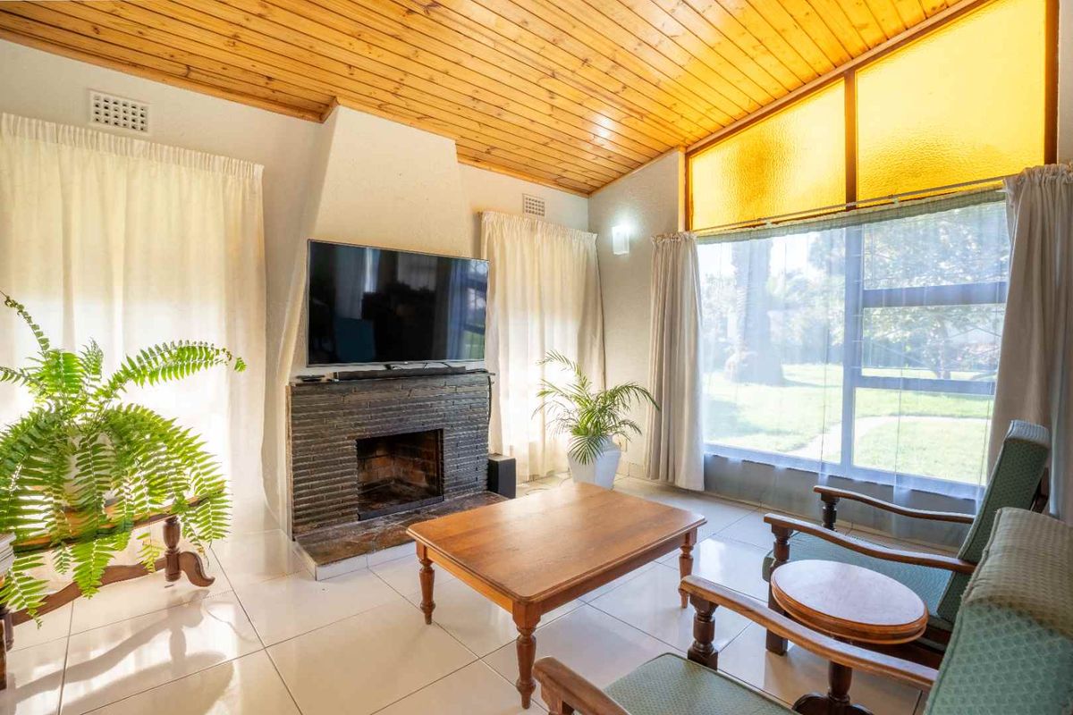 A bright living room featuring a brick fireplace, timber-clad ceiling, and large windows overlooking the garden, finished with neutral tiled flooring.