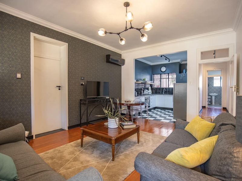 Open-plan living area featuring hardwood floors, decorative wallpaper, and a modern chandelier, seamlessly connecting to a functional kitchen with checkerboard tiling.
