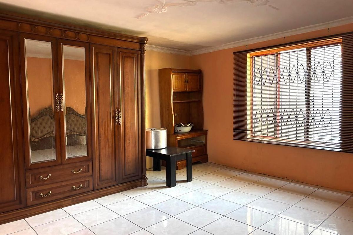 A bedroom featuring tiled flooring, a large built-in wooden wardrobe, and a window fitted with security bars and blinds.