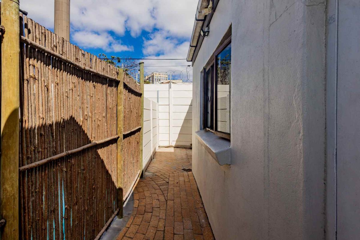 A narrow exterior side passage featuring paved flooring, a boundary fence, and a white rendered building wall with a window.
