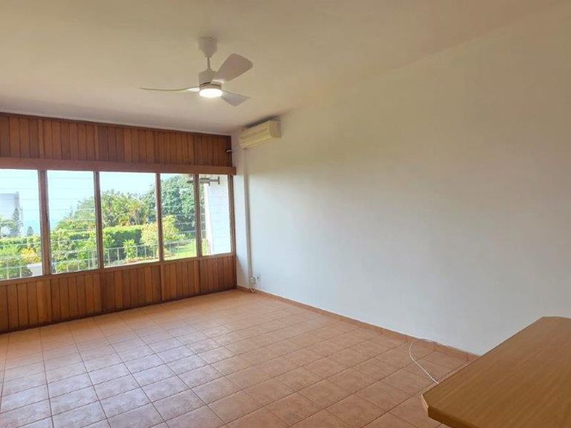 Spacious, light-filled living area featuring tiled flooring, a ceiling fan, split-system air conditioning, and large timber-framed windows with garden views.