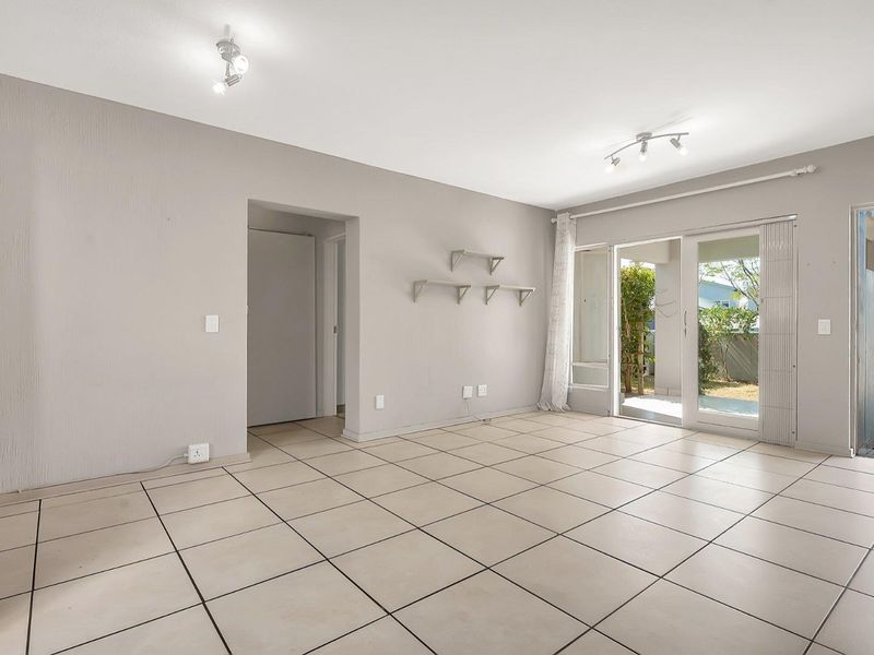 A bright, empty living room featuring neutral tiled flooring, wall-mounted shelving, and a sliding glass door providing access to the outdoor area.