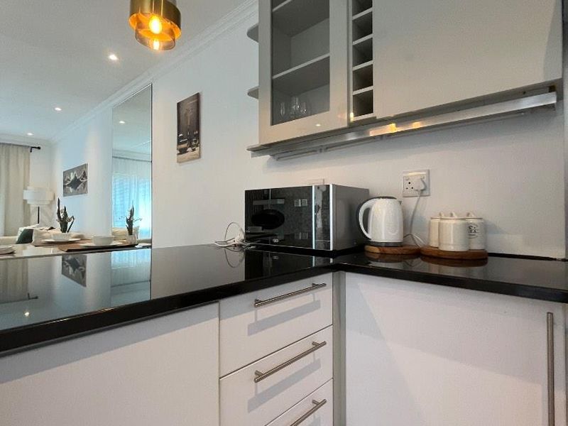 Modern kitchen featuring sleek white cabinetry, black granite countertops, and glass-fronted upper storage, seamlessly flowing into the adjacent living area.