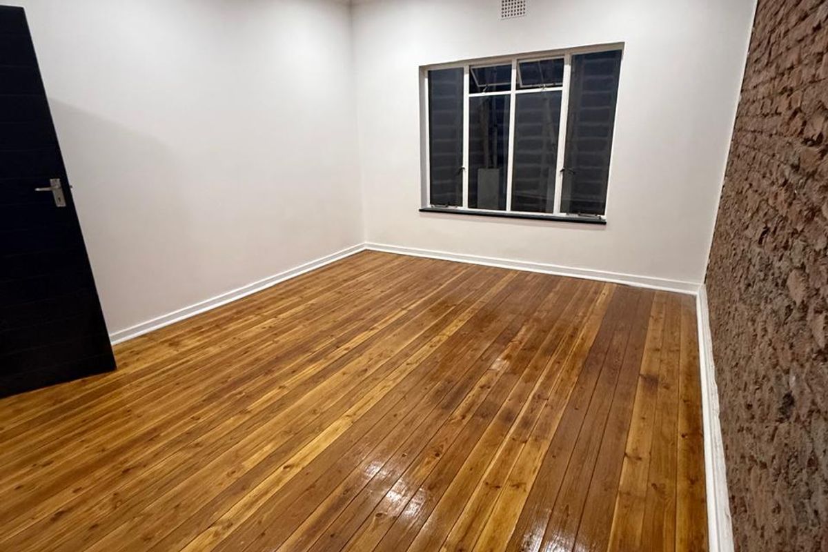 A bright, empty bedroom featuring polished timber flooring, an exposed brick feature wall, and recessed downlighting.