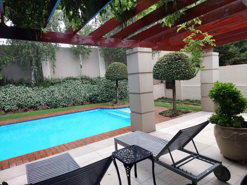 A well-maintained outdoor pool area featuring a covered patio with a timber pergola, paved surround, and established garden landscaping.