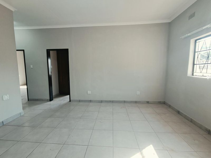 A spacious, empty living area featuring neutral tiled flooring, light grey walls, and a window fitted with security burglar bars.