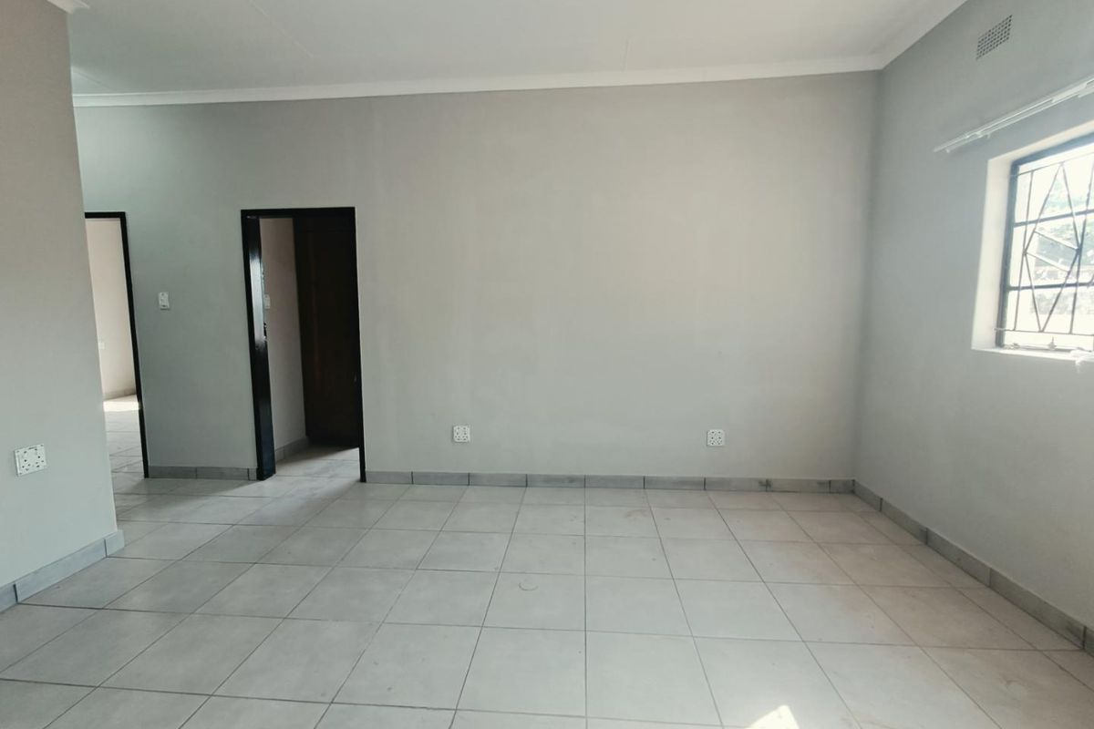 A spacious, empty living area featuring neutral tiled flooring, light grey walls, and a window fitted with security burglar bars.
