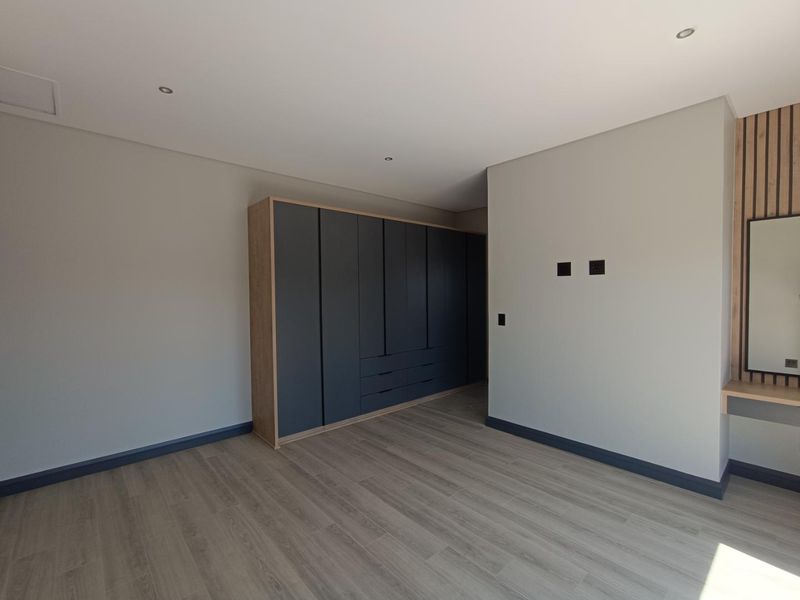 A modern, empty bedroom featuring sleek dark built-in cupboards, light wood-look laminate flooring, recessed lighting, and a dedicated vanity area.