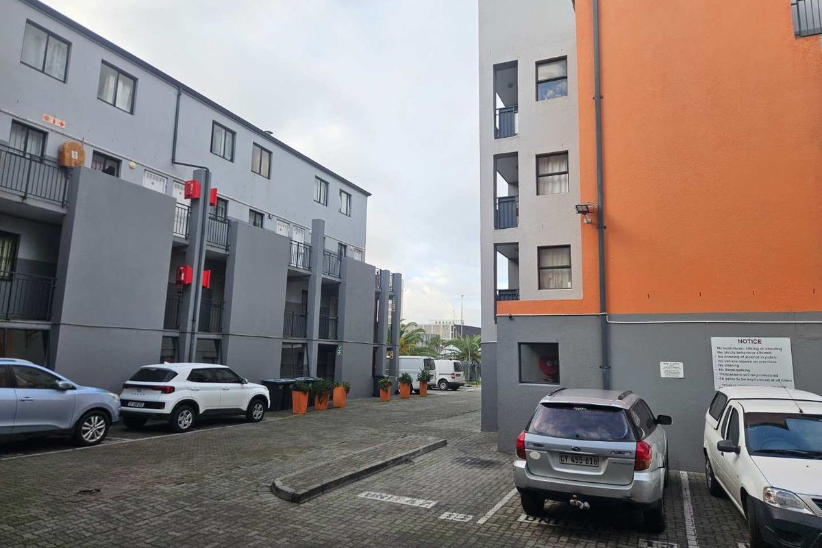 Exterior view of the apartment complex parking area, featuring paved ground, multi-story building facades, and designated parking bays.