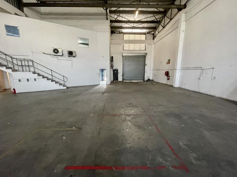Spacious industrial warehouse unit featuring high ceilings with exposed steel trusses, concrete flooring, a large roller shutter door, and an internal staircase.