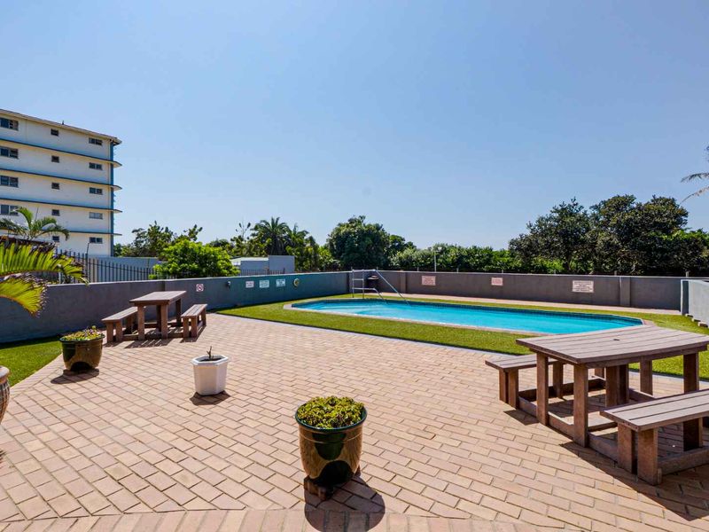 Communal outdoor pool area featuring a paved patio, artificial grass surround, and perimeter walling within an apartment complex.
