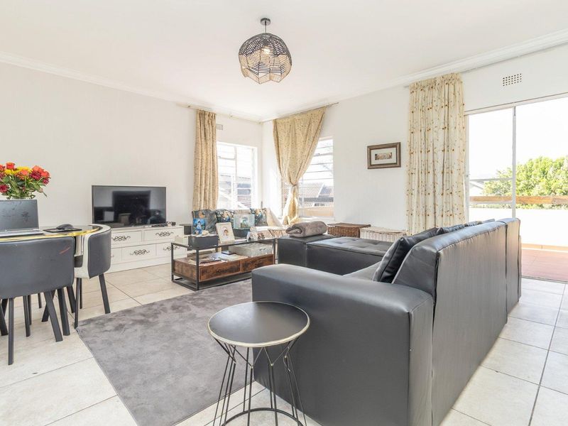A bright, open-plan living area featuring tiled flooring, large windows, and a sliding door providing access to an outdoor balcony space.