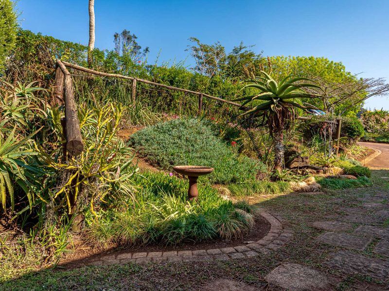 A well-maintained garden area featuring stone paved pathways, established indigenous planting, and rustic timber pole fencing.