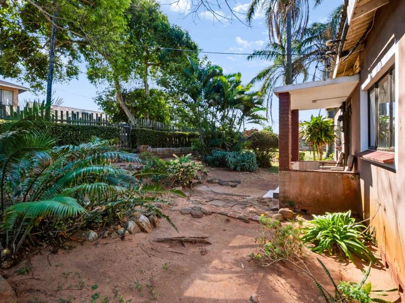A rear exterior view featuring a covered patio area, established garden landscaping, and brick structural elements requiring maintenance.