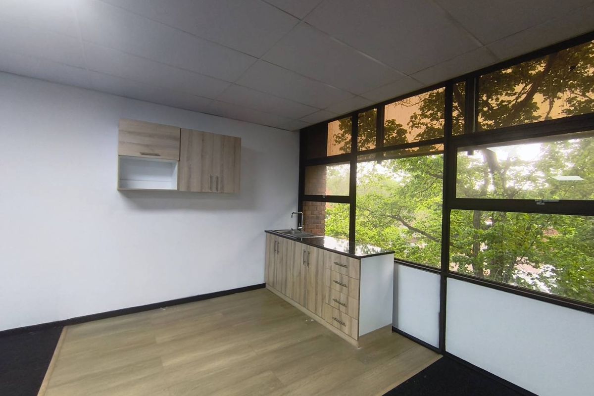 A clean, modern office kitchenette featuring light wood-finish cabinetry, a stainless steel sink, and large windows providing ample natural light and a green outlook.