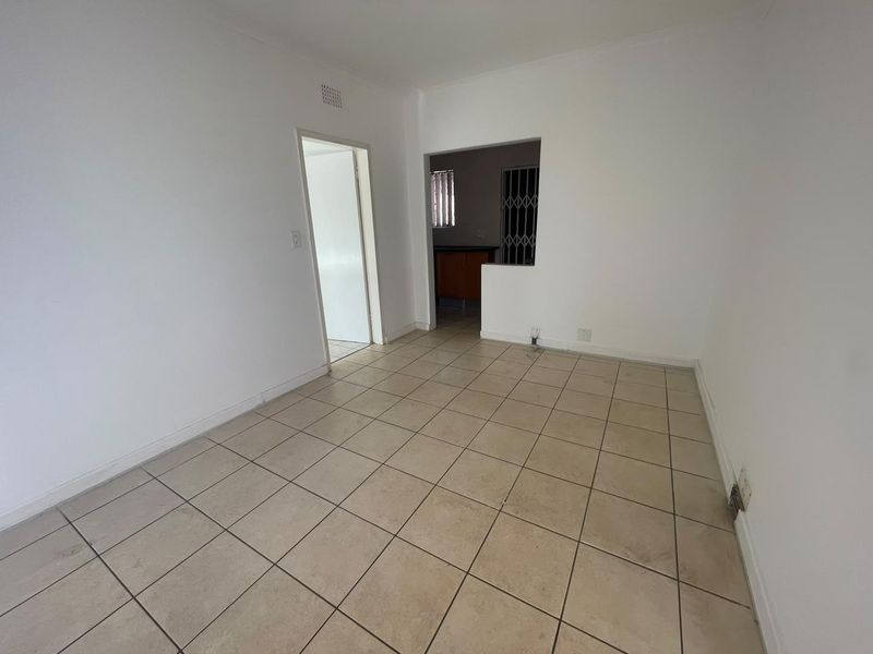 An empty, tiled living area with neutral walls, featuring a pass-through opening to the kitchen and access to an adjacent room.
