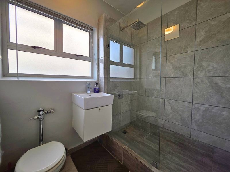 Modern bathroom featuring a glass-enclosed shower with rain shower head, wall-mounted vanity, and neutral grey tiling throughout.