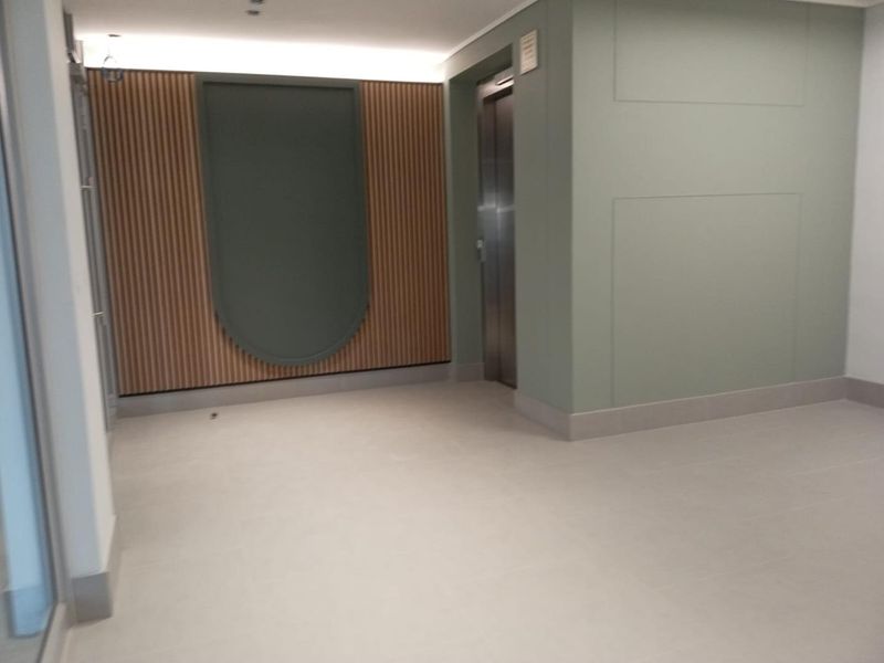 A modern, minimalist hallway featuring a stainless steel elevator, decorative slatted timber wall paneling, and clean neutral floor tiling.