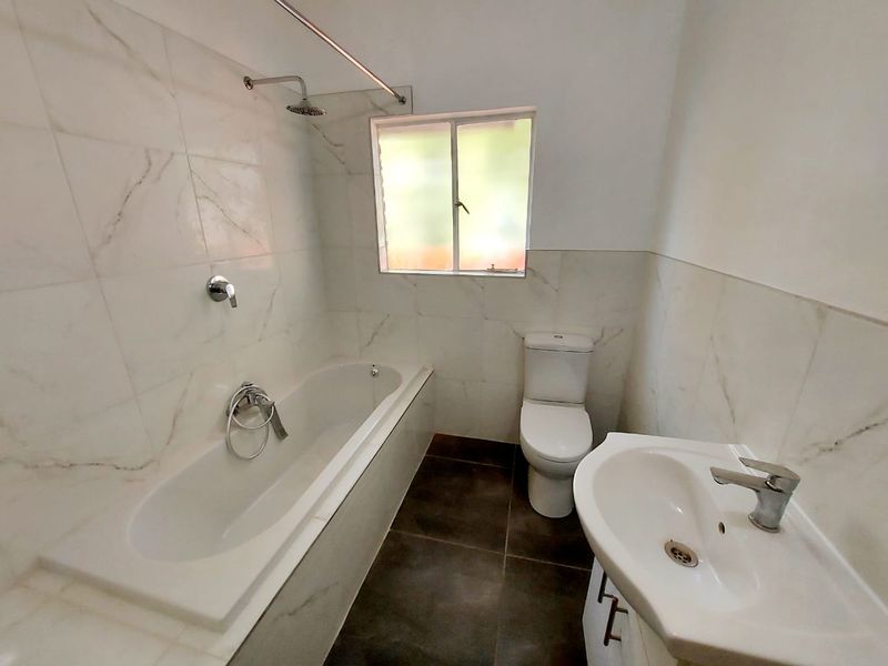 A clean, modern bathroom featuring marble-look wall tiles, a built-in bathtub with shower, a pedestal vanity basin, and a toilet.
