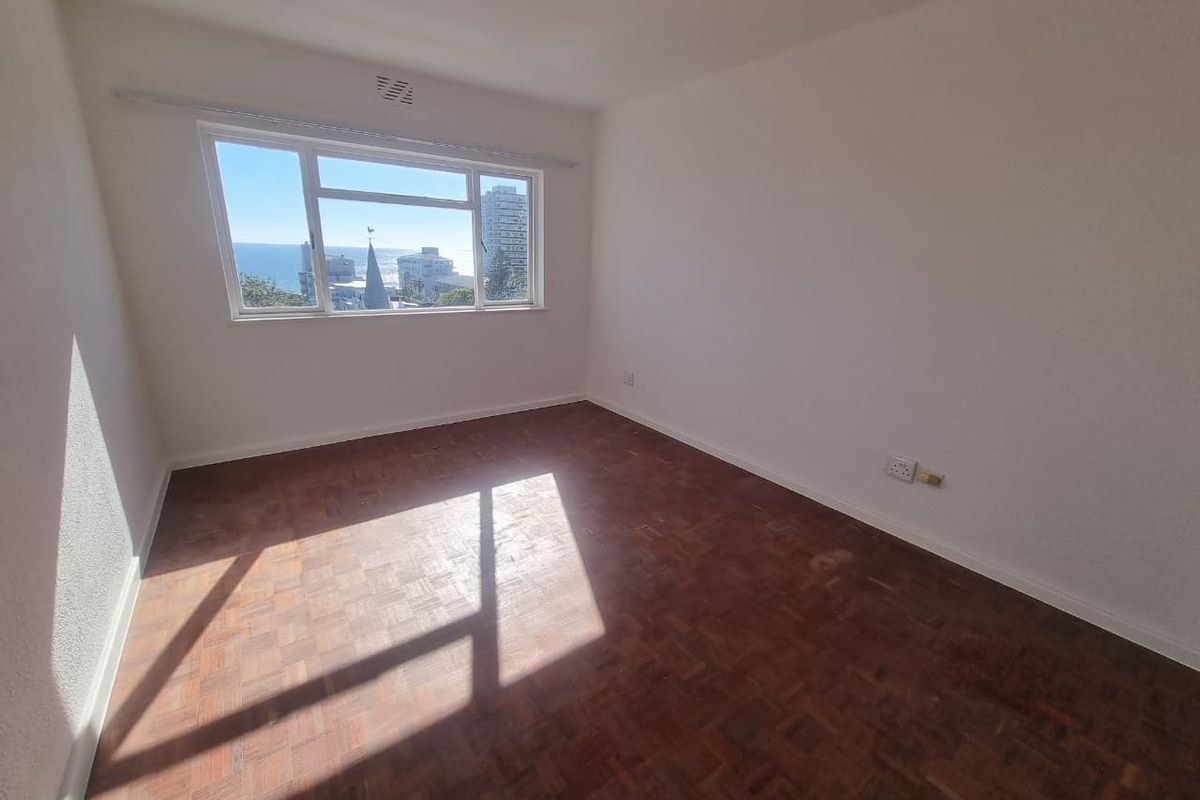 An empty bedroom featuring classic parquet flooring, white walls, and a large window offering a scenic coastal view.
