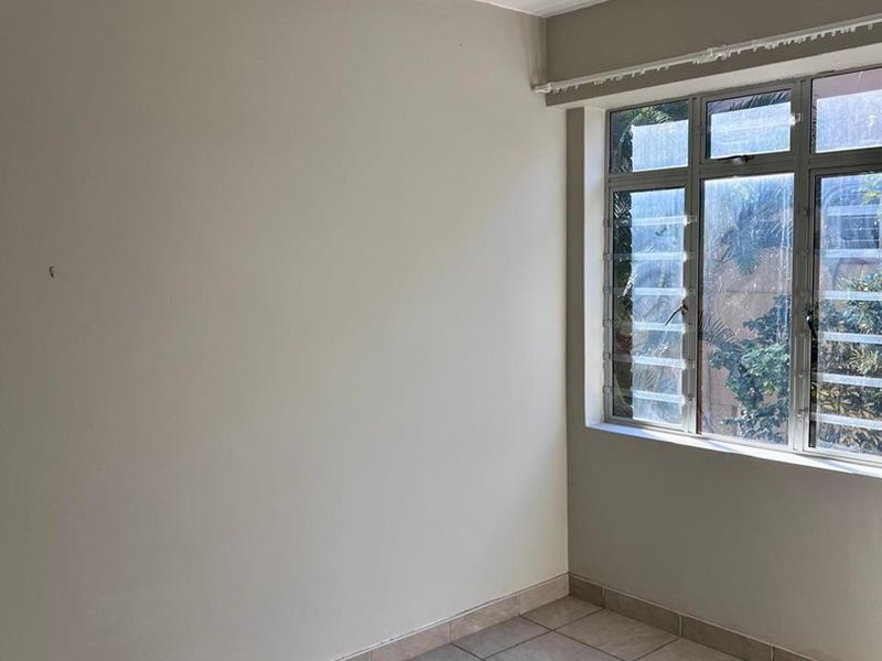An empty bedroom featuring neutral wall tones, tiled flooring, and a ceiling fan, with natural light provided by a window with security bars.