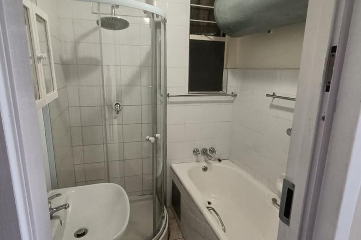 Compact bathroom featuring a shower enclosure, bathtub, and pedestal basin, with an exposed hot water cylinder and dated finishes throughout.